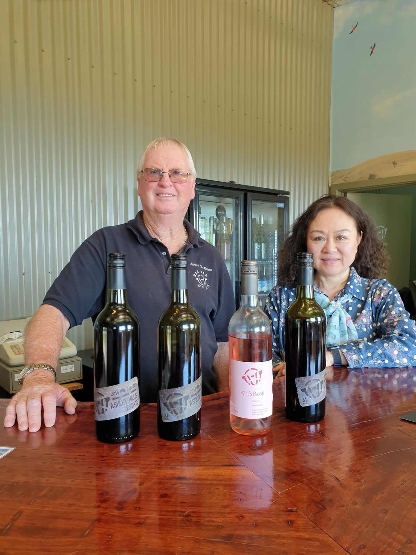 Paul and Wei behind the cellar door bar with bottles of Ashley Hills wine